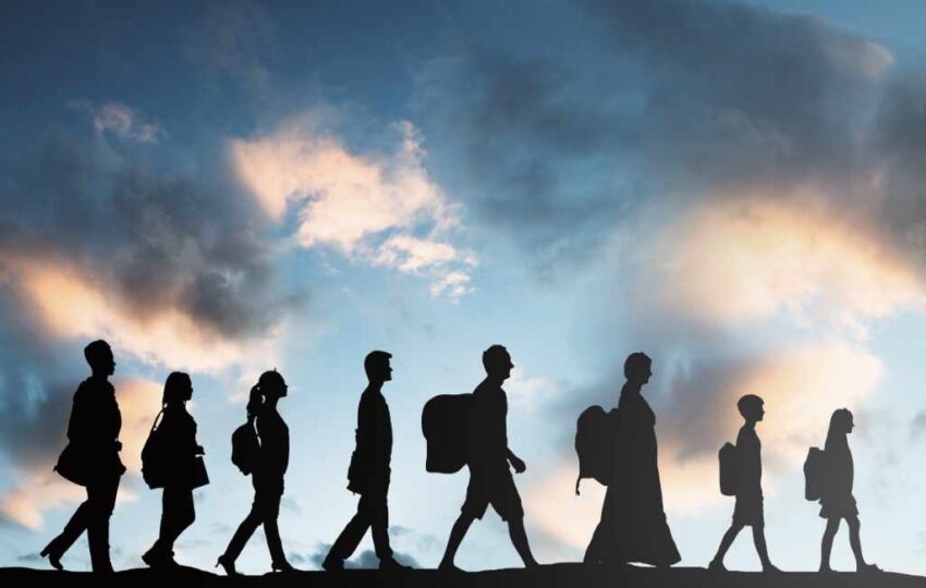 The image seven refugees followinf each other in a queue, making the arduous journey from their homes country to a safer land. Image has silhouettes of individuals of all age, walking from left to right. At the backdrop is a dusky cloudy sky.